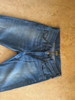 Blå jeans från Lee med raka ben - Säljer ett par klassiska blå jeans från Lee i en snygg tvätt. Jeansen har raka ben, fem fickor och Lee-logga på bakfickan samt ett brunt lädermärke i midjan. Även slitna detaljer för en trendig look. Midjan=32 cm. Innerbenslängden=70 cm. Skriv för frågor!💕