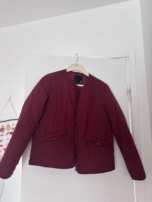 Vinröd pufferjacka - Snygg vinröd pufferjacka med rak passform och rund halsringning. Jackan har två fickor framtill med knappstängning och dragkedja i mitten. Perfekt för kyliga dagar och enkel att matcha med olika outfits.