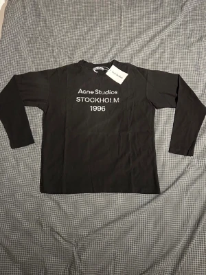 Svart långärmad tröja Acne Studios - Snygg svart långärmad tröja från Acne Studios med tryck 'Acne Studios STOCKHOLM 1996' på bröstet. Klassisk rak passform och rund halsringning. Perfekt till jeans eller kjol för en clean och modern look.