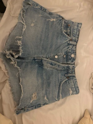 Blå denimshorts med fransar från ZARA - Säljer ett par ljusblå denimshorts från ZARA med fransiga kanter och slitna detaljer. Shortsen har hög midja, klassisk femficksdesign och knappar där fram.