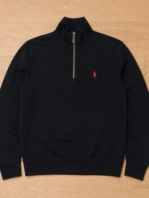 Svart halvzip tröja från Ralph Lauren - Klassisk svart halvzip tröja från Ralph Lauren med röd broderad logga på bröstet. Tröjan har hög krage, ribbade muddar och är tillverkad i mjukt material. Perfekt till jeans eller chinos för en clean look.
