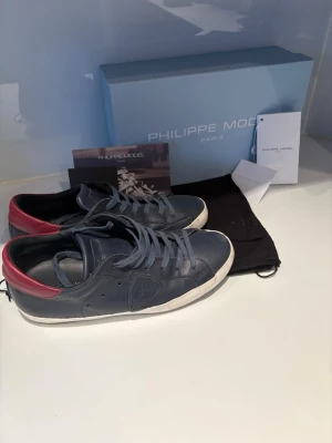Philippe Model blåa sneakers - Snygga sneakers från Philippe Model i blått läder med vita sulor och röda detaljer på hälen. Klassisk låg modell med matchande blå skosnören och diskret logga på sidan. Perfekta för en trendig och avslappnad stil. Mycket bra skick. Storlek 40