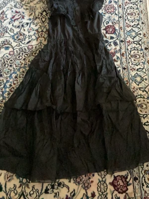 Svart långklänning med volanger - Säljer en svart långklänning med volangdetaljer och knappar framtill. Klänningen har en luftig och flowy passform, perfekt för dig som gillar en elegant och bohemisk stil. Materialet känns lätt och har en snygg struktur.