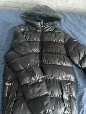 Svart pufferjacka från Alessandro - Snygg svart pufferjacka från Alessandro med glansig finish och huva. Jackan har dragkedjor framtill och på fickorna samt en tydlig logga på bröstet och en patch på ärmen. Perfekt för kalla vinterdagar och riktigt trendig look. FUNKAR M OKSÅ!!
