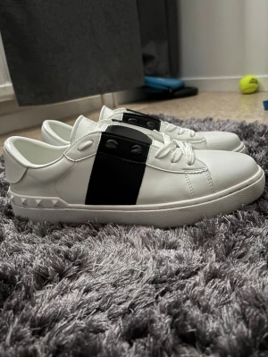 Vita Valentino sneakers med svart detalj - Snygga vita sneakers från Valentino med en bred svart läderdetalj över mitten och klassiska vita skosnören. Skorna har en låg profil och en robust sula med rutmönster. Perfekta för dig som gillar stilrena och exklusiva sneakers.