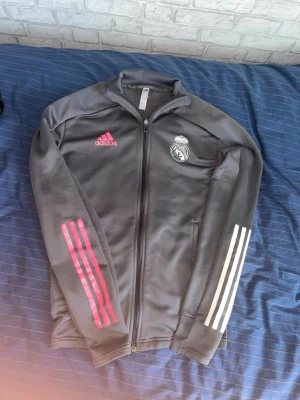 Grå Real Madrid träningsjacka Adidas - Snygg grå träningsjacka från Adidas med Real Madrid-logga på bröstet. Jackan har dragkedja, två fickor och rosa Adidas-logga. Ena ärmen har rosa ränder och den andra vita ränder, vilket ger en unik look. Perfekt för fotbollsträning eller chill.