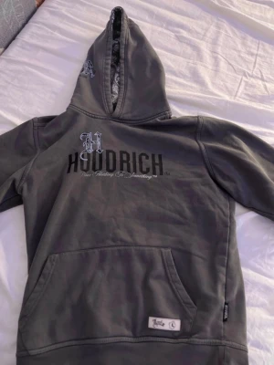 Grå Hoodrich hoodie med tryck - Snygg grå hoodie från Hoodrich med stor logga och text på bröstet samt detaljer på huvan. Klassisk känguruficka framtill och ribbade muddar. Perfekt för en chill och street-inspirerad look.