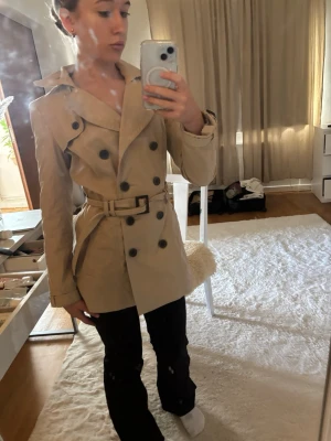 Beige trenchcoat med bälte - Stilren beige trenchcoat med dubbelknäppning och matchande bälte i midjan. Klassisk krage och axelklaffar ger en tidlös look. Jackan har svarta knappar och är tillverkad i ett slätt tyg som känns lätt men ändå skyddande. Perfekt för vår och höst. Vintage Zara 