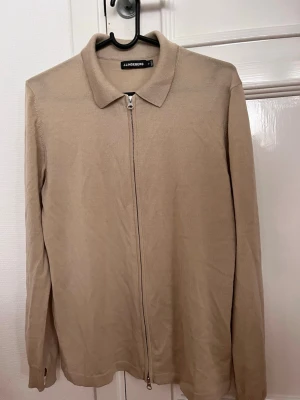 Beige ziptröja från J.Lindeberg - Stilren beige ziptröja från J.Lindeberg med klassisk krage och långa ärmar. Tröjan har en hel dragkedja framtill och är tillverkad i ett tunt, mjukt material som passar perfekt för lager på lager. Enkel och snygg design som funkar till det mesta.