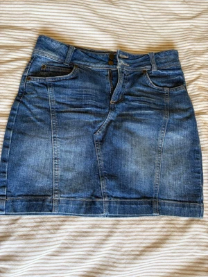 Blå jeanskjol med fickor - Säljer en klassisk blå jeanskjol i kort modell med fem fickor, bälteshällor och knappgylf. Kjolen har en snygg tvättad look och passar perfekt till en avslappnad stil. Materialet är slitstarkt denim och kjolen har en normal passform.