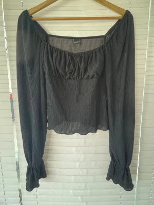 Svart offshoulder blus med långa ärmar - Så himla söt Trendig svart offshoulder blus i ett tunt, lätt och lite transparent tyg. Blusen har långa puffiga ärmar med resår och volang vid ärmslut samt rynkad detalj vid bysten. Perfekt för dig som gillar en romantisk och avslappnad stil.