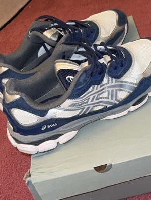 Asics vita och blå sneakers - Snygga sneakers från Asics i vitt och marinblått med grå detaljer. Ovandelen är i mesh och syntet för bra andningsförmåga och komfort. Klassisk Asics-logga på sidan och vadderad häl för extra support. Perfekta för en sportig och trendig look.