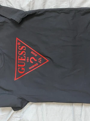 Svart t-shirt från Guess med röd logga - Svart t-shirt från Guess med den klassiska röda triangelloggan på bröstet. T-shirten har rund hals och korta ärmar. Perfekt för dig som gillar stilrena plagg med tydlig branding.