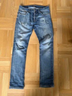 Blå slitna jeans från Nudie Jeans - Säljer ett par blå jeans från Nudie Jeans med slitningar och distressed detaljer på låret. Jeansen har klassisk femficksmodell, slimmade  ben och är tillverkade i denim. Perfekta för en avslappnad och trendig look.