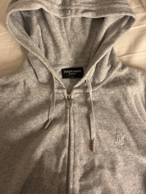 Grå Juicy Couture zip hoodie - Säljer en ljusgrå hoodie från Juicy Couture i storlek XS. Hoodien har dragkedja, huva med dragsko och silverfärgade detaljer på snörena. Broderad JC-logga på bröstet. Mjuk och skön bomullsblandning, perfekt för en chill och avslappnad stil. Säljer hela setet för 1200kr och byxorna är i xxs medans zip hoodien är i xs.