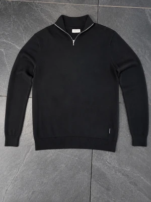 Svart halvzip-tröja från Lindbergh - Snygg svart tröja från Lindbergh med halvzip och ståkrage. Slim fit-modell i mjukt material, perfekt för lager på lager. Ribbstickade muddar vid ärmslut och nederkant ger en clean look. Passar till både jeans och chinos.