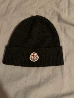 Svart Moncler mössa i ull - Säljer en svart ribbstickad mössa från Moncler med uppvikt kant och klassisk Moncler-logga framtill. Mössan är gjord i mjuk ull och har en enkel, stilren design som passar perfekt till kalla dagar.