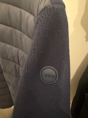 Mörkblå cardigan från Colmar - Snygg mörkblå cardigan från Colmar med vadderad huva och ärm med Colmar-logga. Tillverkad i ett stickat material som ger en cool och sportig vibe. Perfekt för dig som gillar streetwear och vill ha något bekvämt och stilrent.