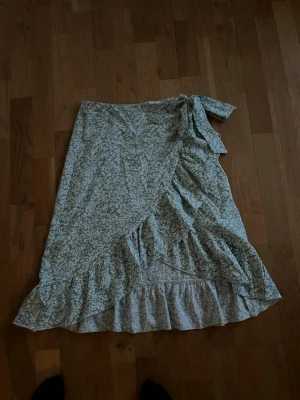 Grön blommig omlottkjol från SHEIN - Säljer en grön blommig omlottkjol från SHEIN med volangkant och knyt i midjan. Kjolen har ett romantiskt blommönster och är perfekt för vår och sommar. Lätt och luftig modell som ger en avslappnad men söt look.