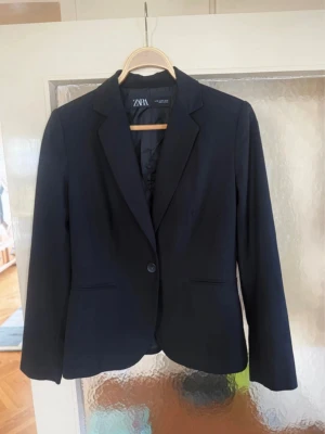Svart kavaj från Zara - Stilren svart kavaj från Zara med klassisk krage och en knapp framtill. Kavajen har två fickor och är helfodrad för extra komfort. Perfekt för en clean och snygg look. Passar till många olika outfits och är enkel att matcha.
