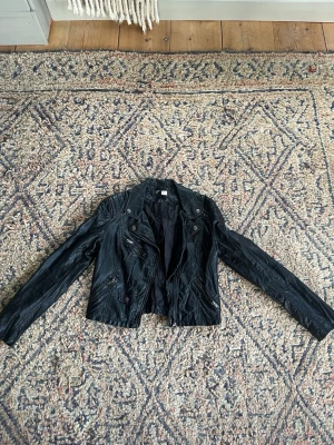 Svart biker skinnjacka med dragkedjor - Snygg svart skinnjacka i bikerstil med flera dragkedjor och detaljer på axlarna. Jackan har lång ärm, snedställd dragkedja framtill och två fickor med dragkedja. Materialet är glansigt svart skinn som ger en cool look. Lädret på kragen är något sprucket som man kan se på bild