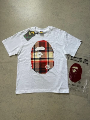 Vit Bape t-shirt  - Vit t-shirt från Bape med klassisk ap-logo i röd, beige och svart rutig design på bröstet. T-shirten har rund hals och korta ärmar. Perfekt för dig som gillar streetwear och vill sticka ut med en unik detalj framtill. Size S, helt ny och aldrig använt🤩