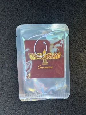 Suryoyo Bildoft - Bildoft med Suryoyo-symbol i guld på vinröd bakgrund. Hänget är fyrkantigt och har ett vitt snöre för upphängning. Kommer i en genomskinlig plastförpackning. Perfekt som dekoration till bilen eller rummet.