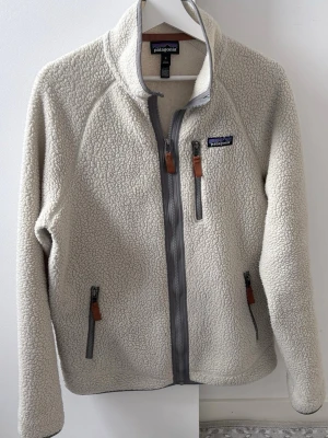 Patagonia fleece jacka  - Säljer en offwhite fleecejacka från Patagonia med grå dragkedjor och bruna detaljer. Jackan har hög krage, tre fickor med dragkedja och ett mjukt, fluffigt material som håller dig varm. Perfekt för kyliga dagar och outdoor-häng.