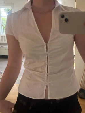 Vit kortärmad skjorta med knappar - Säljer en vit skjorta med korta ärmar och djup v-ringning. Skjortan har figurnära passform och knappar framtill. Perfekt till dig som gillar en clean och enkel stil. Passar S också skulle jag säga 