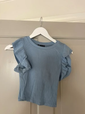 Ljusblå ribbad topp med volangärm - Supersöt ljusblå topp från Zara med ribbad struktur och volangdetaljer på ärmarna. Toppen är ärmlös och har en croppad passform som passar perfekt till höga jeans eller kjol. Enkel att styla för en trendig look.