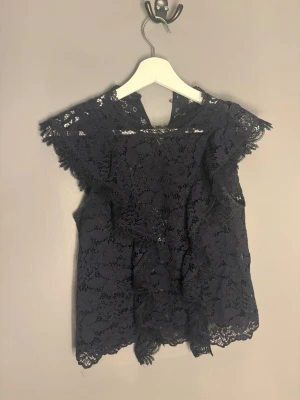 Svart spetsblus med volangdetaljer - Supersnygg svart blus i spets med volangdetaljer och korta ärmar. Perfekt för dig som gillar romantisk stil och vill ha något extra till outfiten. Blusen är genombruten och har fina spetsmönster över hela plagget.
