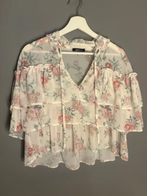 Blommig volangblus - Söt blus från GINATRICOT i tunn, vit mesh med pastellfärgade blommor. Blusen har volanger, v-ringning med knytband. Perfekt för en romantisk och trendig look.