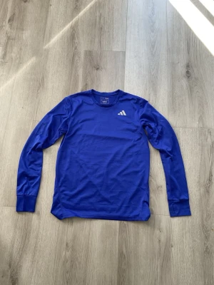 Blå långärmad träningströja Adidas - Snygg blå långärmad träningströja från Adidas med rund hals och diskret logga på bröstet. Tillverkad i ett lätt och funktionellt material som andas, perfekt för träning eller löpning. Enkel och stilren design som passar till det mesta.