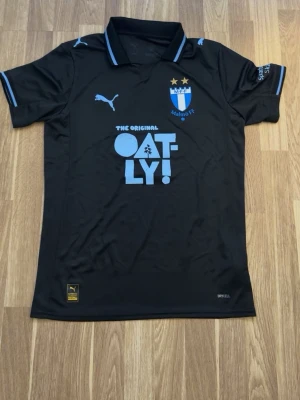 Svart Malmö FF matchtröja Puma - Säljer en svart Malmö FF matchtröja från Puma med ljusblå detaljer och klubbmärke på bröstet. Tröjan har sponsorlogga från Oatly och klassisk krage med ljusblå rand. Tillverkad i funktionsmaterial (Drycell, Re:Fibre) som andas.
