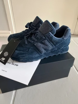 Stone Island x New Balance blå sneakers - Unika sneakers från Stone Island x New Balance i mörkblå mocka. Klassisk låg modell med tydlig N-logga på sidan och Stone Island-märke på plösen. Skorna har matchande blå snören och en robust sula för extra komfort. Perfekta för dig som gillar stilrena och exklusiva samarbeten.