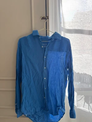 Blå linneskjorta från Massimo Dutti - Snygg blå skjorta från Massimo Dutti i 100% linne. Skjortan har klassisk krage, långa ärmar och vita knappar framtill. Perfekt för en avslappnad men stilren look. Luftigt material som passar bra till varmare dagar.