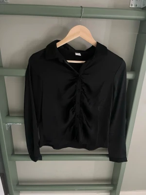Svart satinblus från Lindex - Stilren svart blus i glansigt satinmaterial från Lindex. Klassisk krage, knäppning framtill och långa ärmar. Perfekt för en chic och enkel look. Materialet ger en lyxig känsla och blusen har en lätt rynkning vid knapparna.