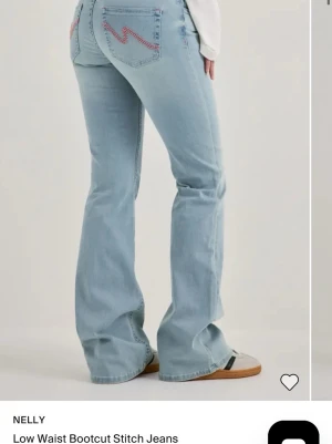 Bootcut ljusblå jeans med zigzag-detalj - Säljer ett par ljusblå bootcut jeans från Nelly med låg midja. Jeansen har röda zigzag-sömmar på bakfickorna och klassisk femficksdesign. Tillverkade i denim med en avslappnad passform och utsvängda ben. Perfekta för en trendig och avslappnad look.