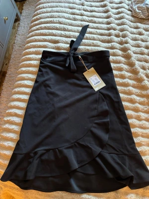 Svart omlottkjol med volang - Snygg svart omlottkjol med volangkant och knytband i midjan. Passar till många olika looks och är enkel att matcha.