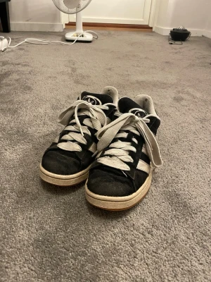Svarta Adidas Campus sneakers - Säljer ett par klassiska Adidas Campus sneakers i svart mocka med vita ränder och vit sula. Skorna har breda vita skosnören och gummisula i brunt. 
