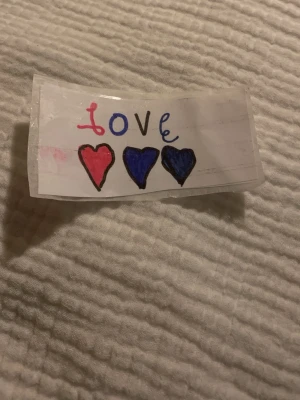 Handgjort dekormärke med hjärtan - Ett handgjort dekormärke i vitt papper, laminerat med tejp. Märket har ordet 'love' i rosa och blått samt tre hjärtan i rött och blått. Perfekt som personlig dekoration till smycken, accessoarer eller pyssel.
