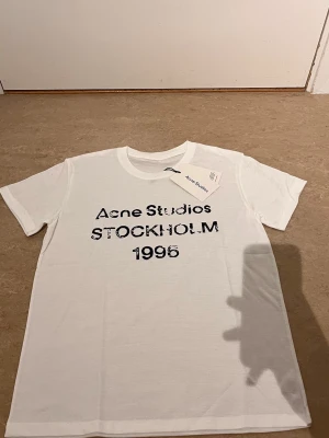 Vit Acne Studios t-shirt med tryck - Vit t-shirt från Acne Studios med svart tryck på bröstet där det står 'Acne Studios STOCKHOLM 1996'. Klassisk rund hals och korta ärmar. Tillverkad i mjuk bomull för en skön och avslappnad look.