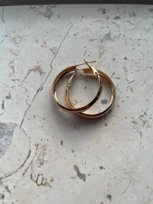  guldfärgade creoler - Säljer ett par  klassiska creoler i guldfärgad metall. Örhängena har en rund form och stängs med ett smidigt klicklås. Perfekta för dig som gillar stilrena och tidlösa accessoarer som ändå syns. Helt nya och oanvända . Obs ! Inte guld, okänt material. 