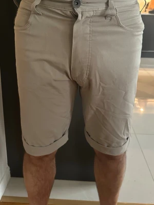 Beige shorts från Armani Jeans - Snygga beige shorts från Armani Jeans med uppvikta benslut och klassisk femficksdesign. Shortsen har bälteshällor, knappgylf och en liten logga på ena bakfickan. Perfekta för en avslappnad och stilren look.