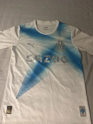 Fotbollströja från marseille - Säljer en vit Marseille matchtröja från Puma med blått grafiskt mönster över bröstet. Tröjan har klubbmärke, Puma-logga och Cazoo-tryck framtill. Tillverkad i ett lätt och ventilerande material, perfekt för fotboll eller träning.