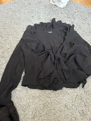 Svart omlottblus från Gina Tricot - Snygg svart omlottblus från Gina Tricot i storlek XS. Blusen har långa ärmar, volangkantad krage och knytband framtill. Materialet är lätt skrynkligt och ger en trendig look. Perfekt att styla med jeans eller kjol för en chic outfit.