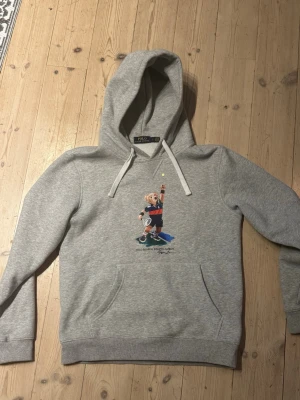 Grå hoodie Polo Bear Ralph Lauren - Säljer en grå hoodie från Polo Ralph Lauren med den ikoniska Polo Bear-trycket framtill. Hoodien har huva med vita dragsnören och en klassisk känguruficka. Tillverkad i mjuk bomullsblandning och har ribbade muddar vid ärmar och nederkant. Storlek L men sitter mer som M.