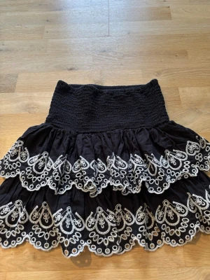Svart kortkjol med broderad spets - Supersöt svart kortkjol med bred smock i midjan och dubbla volanger. Kjolen har vit broderad spetsdetalj längs kanterna som ger en romantisk vibe. Perfekt att styla med en enkel topp eller hoodie. Skulle säga att det passar xxs/xs också.