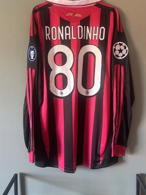 AC Milan Ronaldinho långärmad tröja - Säljer en långärmad AC Milan matchtröja från Adidas med Ronaldinho och nummer 80 på ryggen. Tröjan har röda och svarta ränder, Champions League-patchar på ärmarna och Bwin som sponsor på bröstet. Perfekt för fotbollsfans!