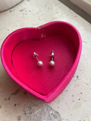 Silverfärgade färger glittriga kulörhängen - Säljer ett par små silverfärgade  örhängen med glittrande kulor. Örhängena är i metall och har en enkel, stilren design som passar till många olika looks.. Perfekta för dig som gillar diskreta men snygga accessoarer. OBS!  inte äkta silver och boxen ingår ej! Nya och oanvända. 